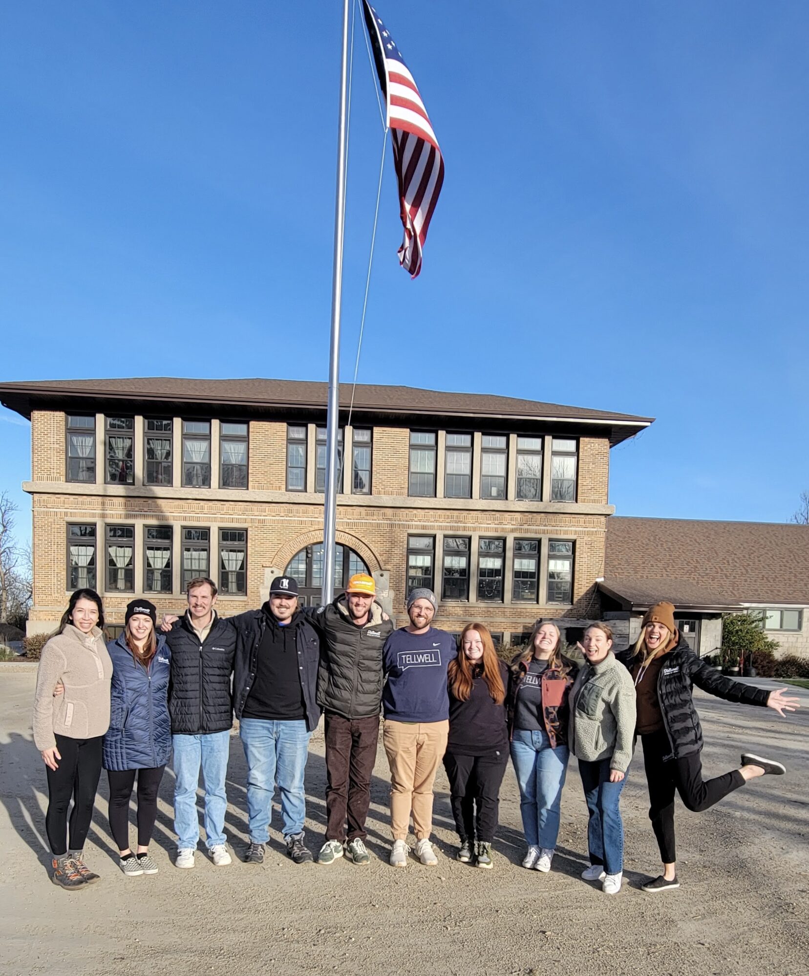A group of nine people, smiling and arm in arm, stand in front of a brick building under a clear blue sky with an American flag waving above.