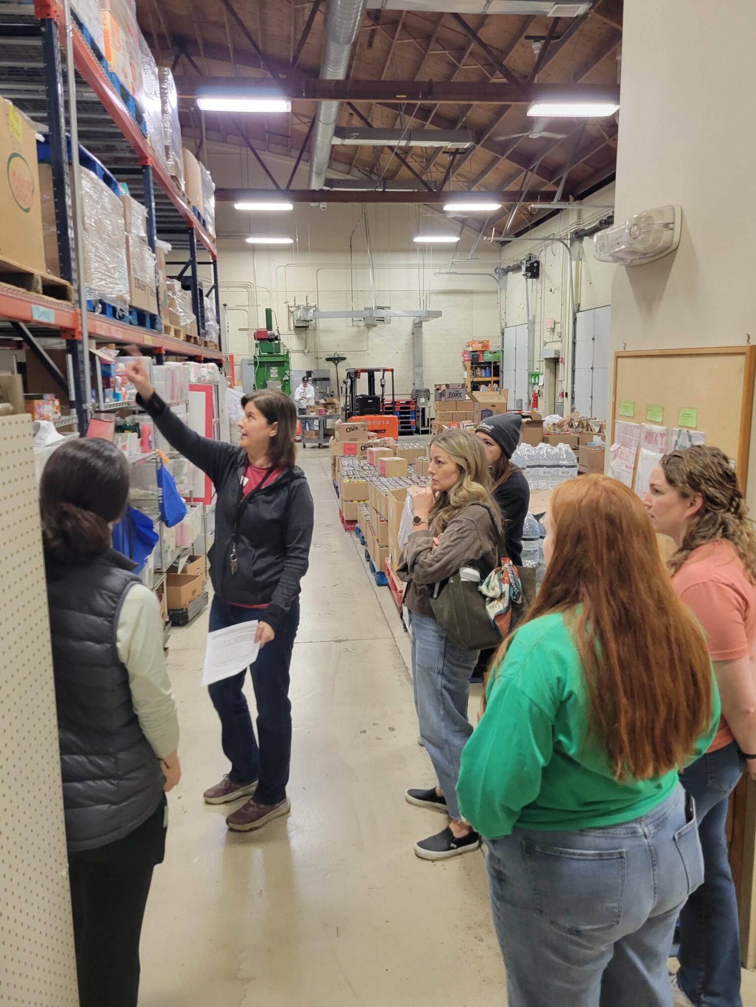 A group of people stand in a warehouse aisle while a woman gestures toward items on a high shelf during a tour or demonstration. Boxes and supplies are stacked on industrial racks, and the group listens attentively in the spacious, well-lit storage area.