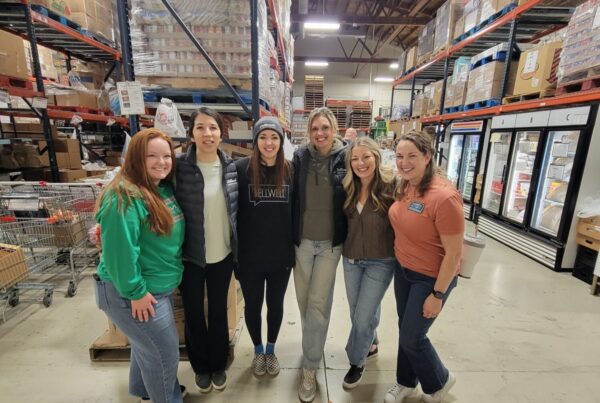 A group of six women stand for a photo inside a warehouse. They are in a food pantry and appear glad to be volunteering.