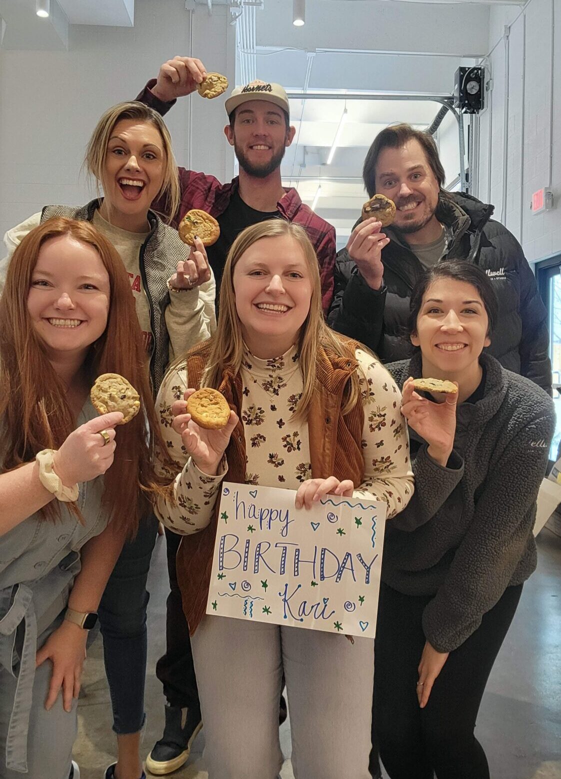 A group of six people smile for a photo. They are all holding cookies and the woman in the front is holding a sign that says “Happy birthday Kari!”