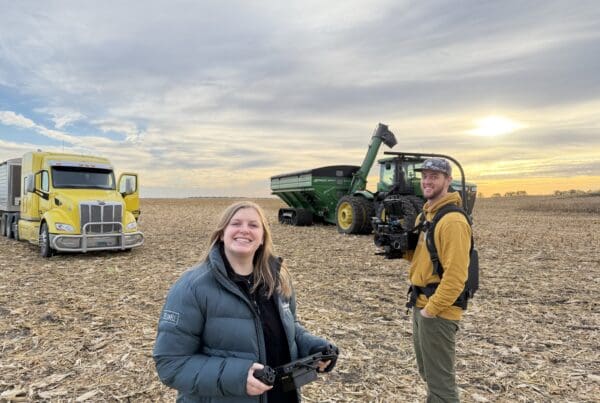 Two people standing in a harvested cornfield at sunrise, one holding a camera monitor and the other carrying a camera rig. A yellow semi-truck and green tractor are parked nearby under a partly cloudy sky.