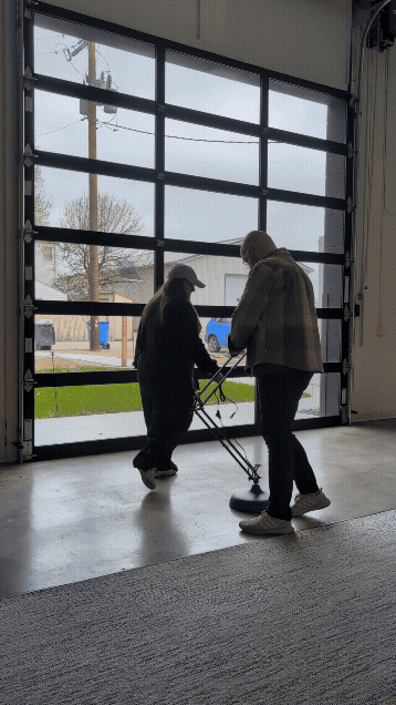 Two people stand inside a garage-style space with large glass doors, working together to attach a tall lamp to its base. They spin the lamp around playfully while securing it, with light coming through the overcast scene outside.
