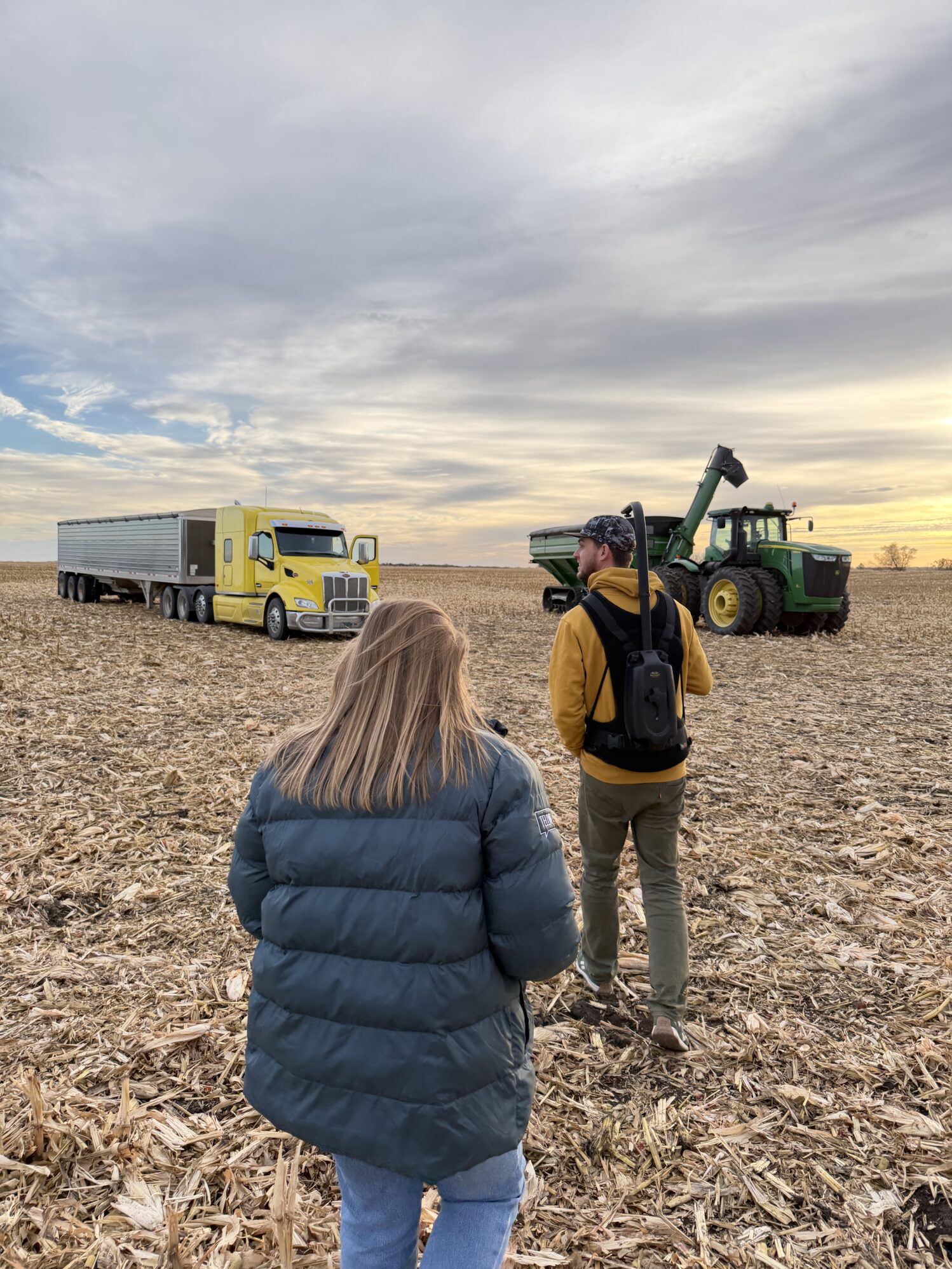 Two people walk through a harvested cornfield toward a yellow semi-truck and a green tractor under a cloudy sky at sunrise. One person wears a dark puffer jacket, and the other wears a mustard hoodie and carries camera gear on their back.