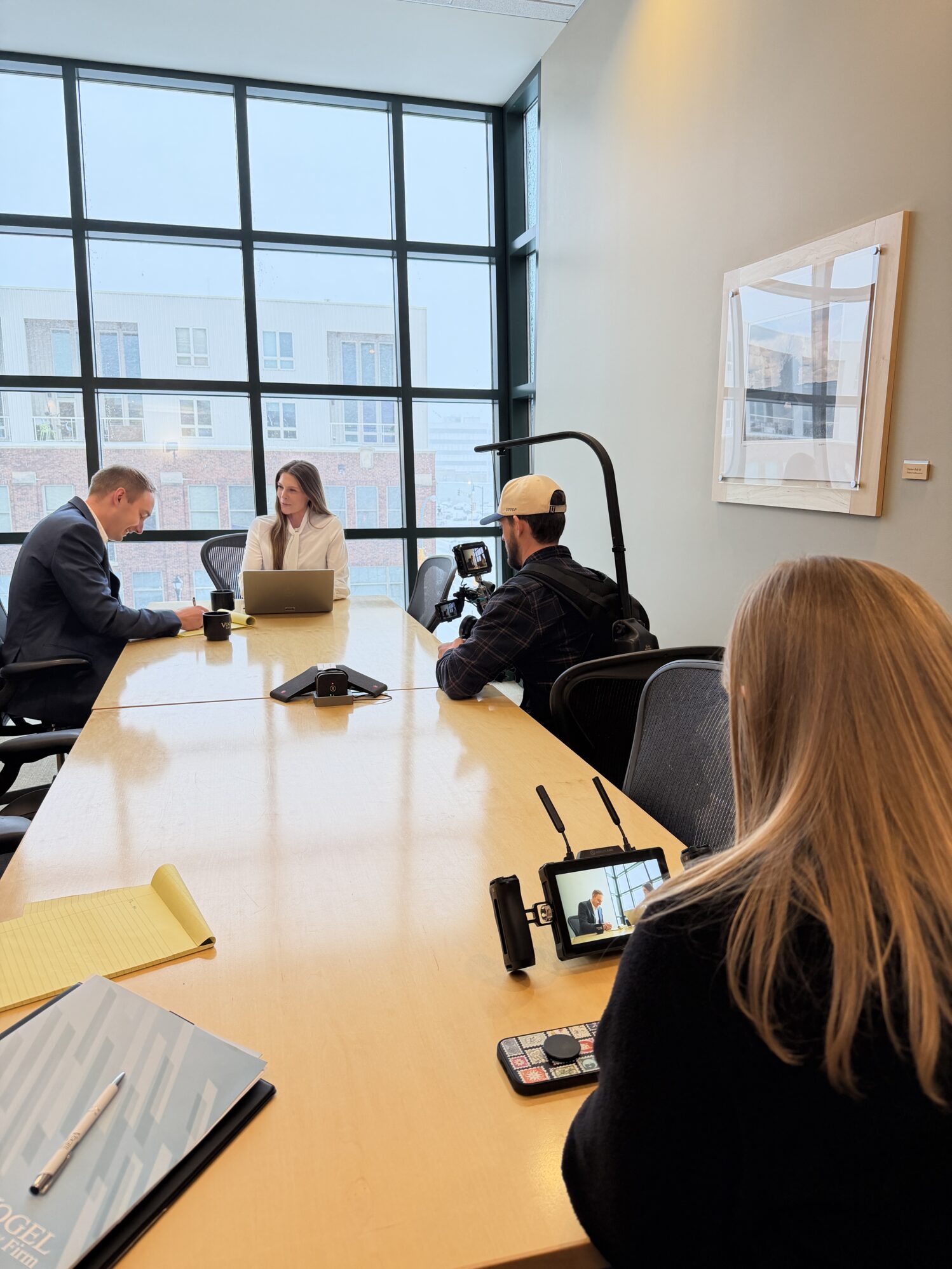 A man operates a camera while a woman watches on a monitor. Two people are being filmed at a conference table, the man is taking notes on a legal pad and the woman is using a laptop. There are large windows behind them and it appears to be snowing outside.