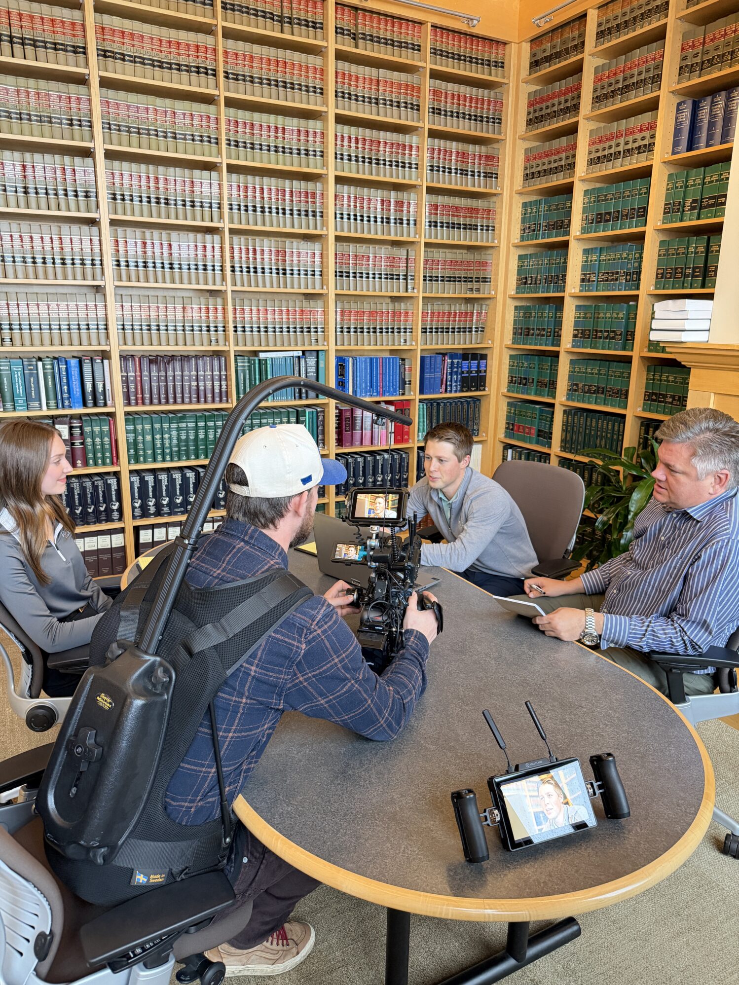 A man operates a camera and films a group of three people sitting at a table. They are in a library with tall shelves filled with legal books. There are two men sitting on one side of the table, and one woman on the other side. They are having a discussion and taking notes on notebooks and laptops.