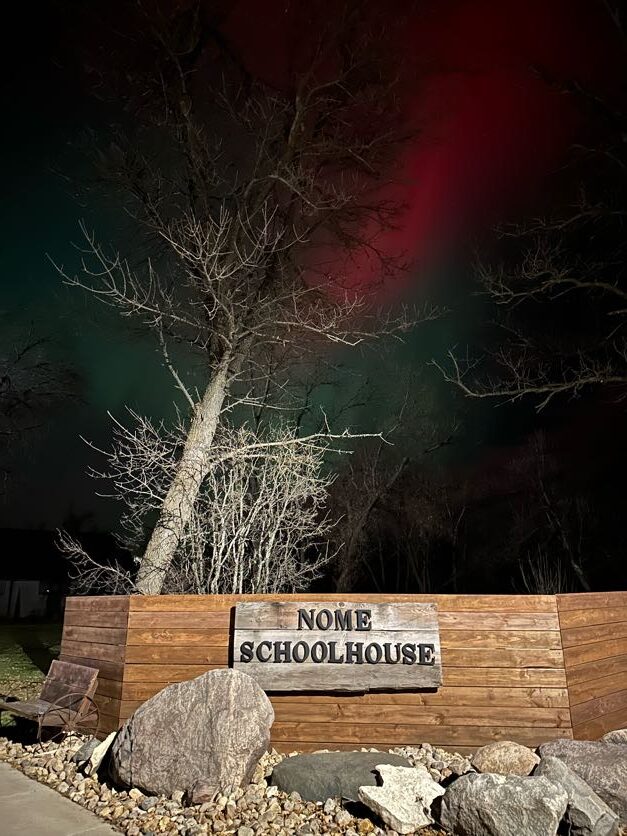 Night scene with silhouetted leafless trees under a green and red aurora. A wooden sign reads “Nome Schoolhouse,” surrounded by rocks.