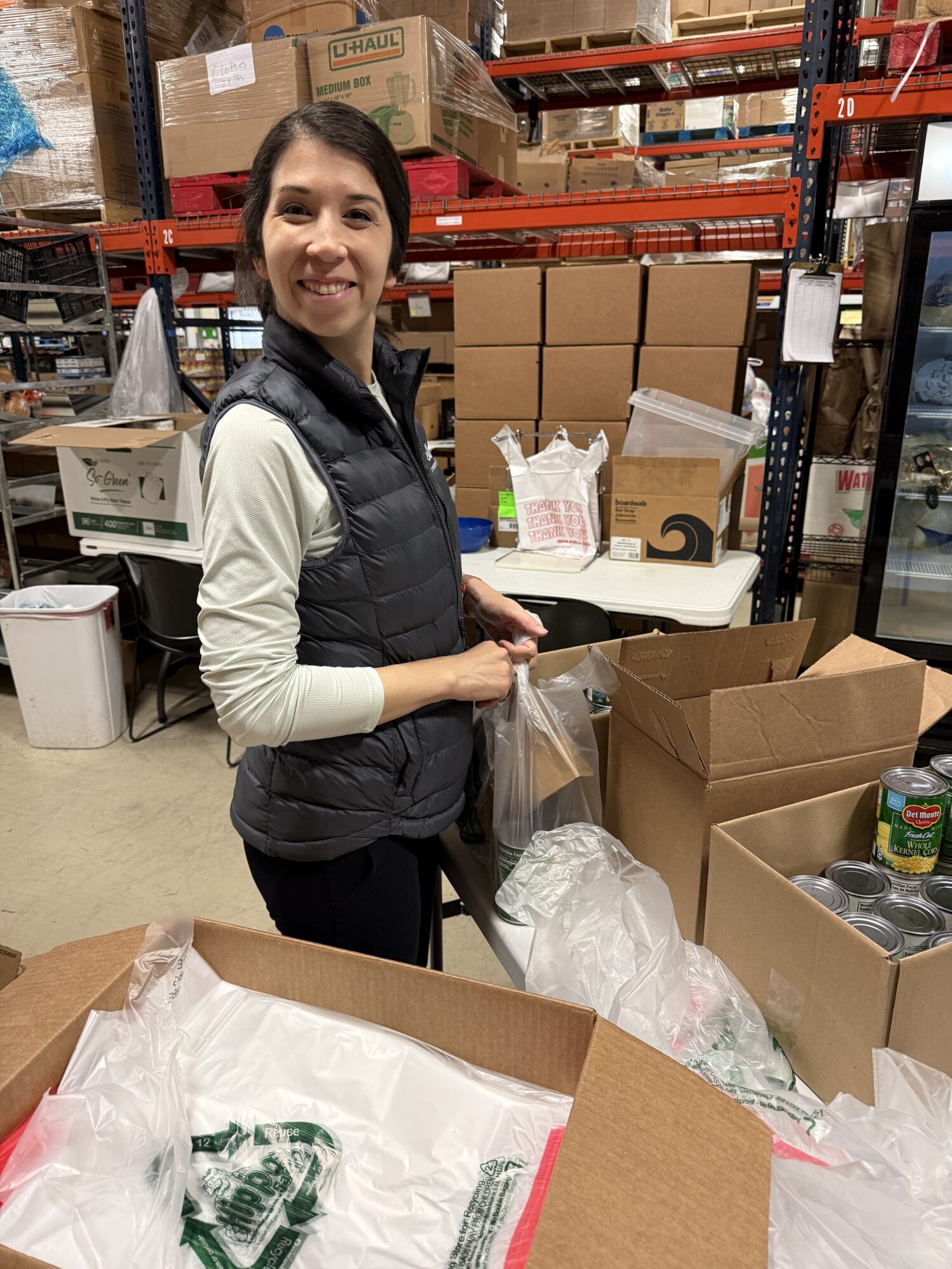 A woman smiles at the camera as she is bagging groceries. She is in a warehouse filled with cardboard boxes which appears to be a food pantry.