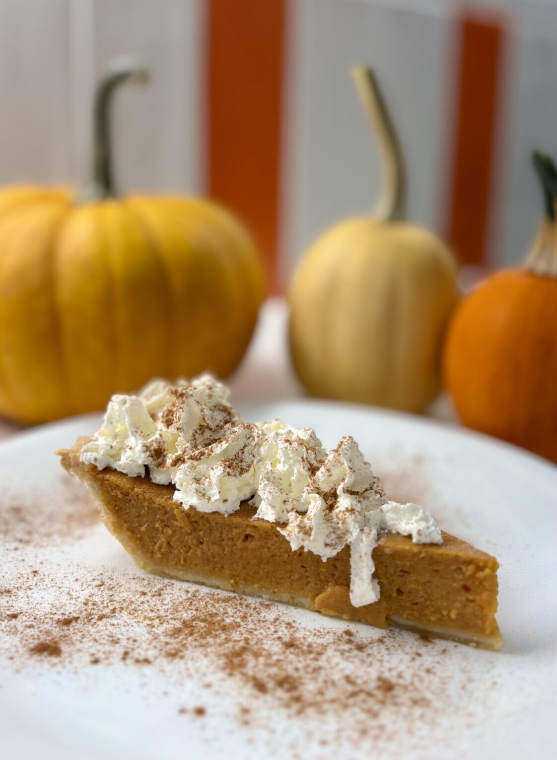 A piece of pumpkin pie with whipped cream and cinnamon sits on a white plate. There are three pumpkins on the background along with a white and orange tiled wall.