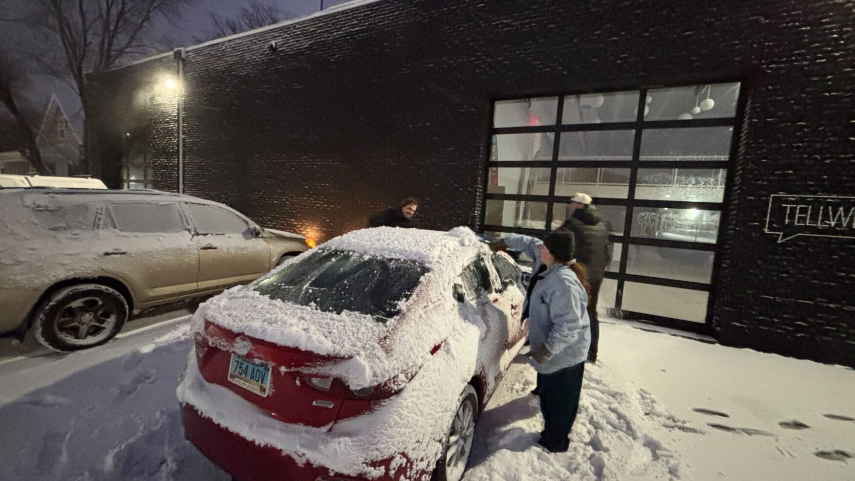 A group of three people stand around a red car, scraping snow and ice off the windows. There is snow on the ground and falling. They are standing in front off a large black building with a "Tellwell" sign.