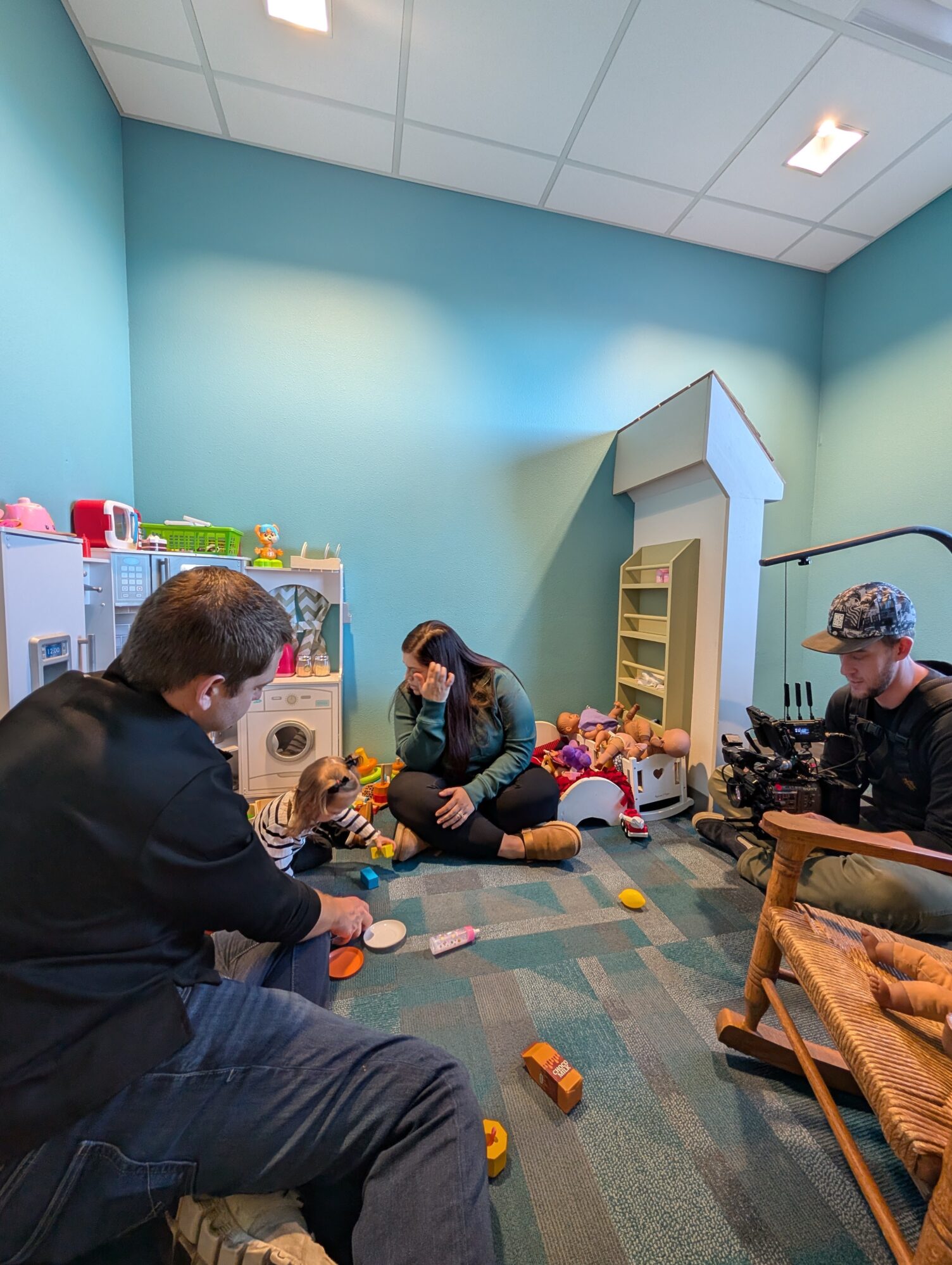 A small group in a playroom setting, including two adults sitting on the floor surrounded by toys and a child playing with them. A cameraman is filming the interaction.