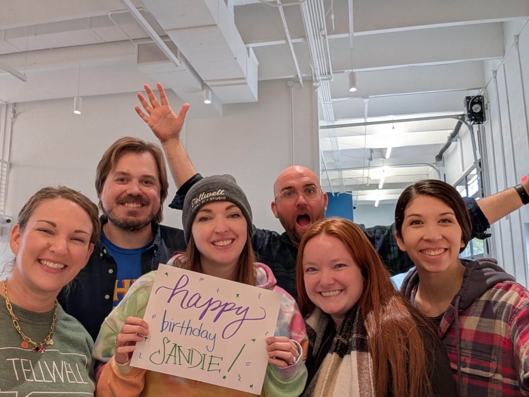 Six people smiling and posing indoors, holding a colorful handwritten sign that says “Happy Birthday Sandie!” One person in the back has their arms raised excitedly.