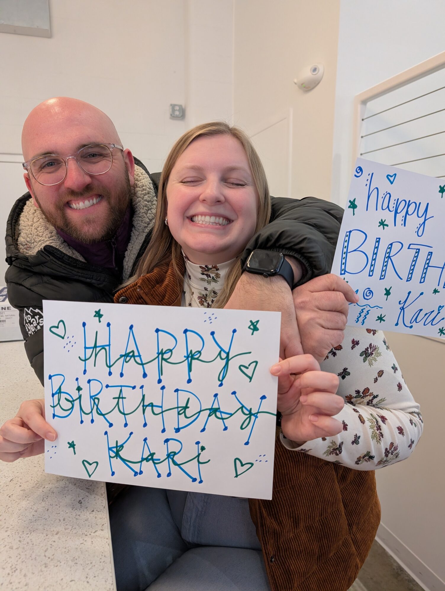 A man and a woman pose for a photo. They are both holding signs that read “Happy birthday Kari!” The man has his arms wrapped around the woman and they are both smiling.