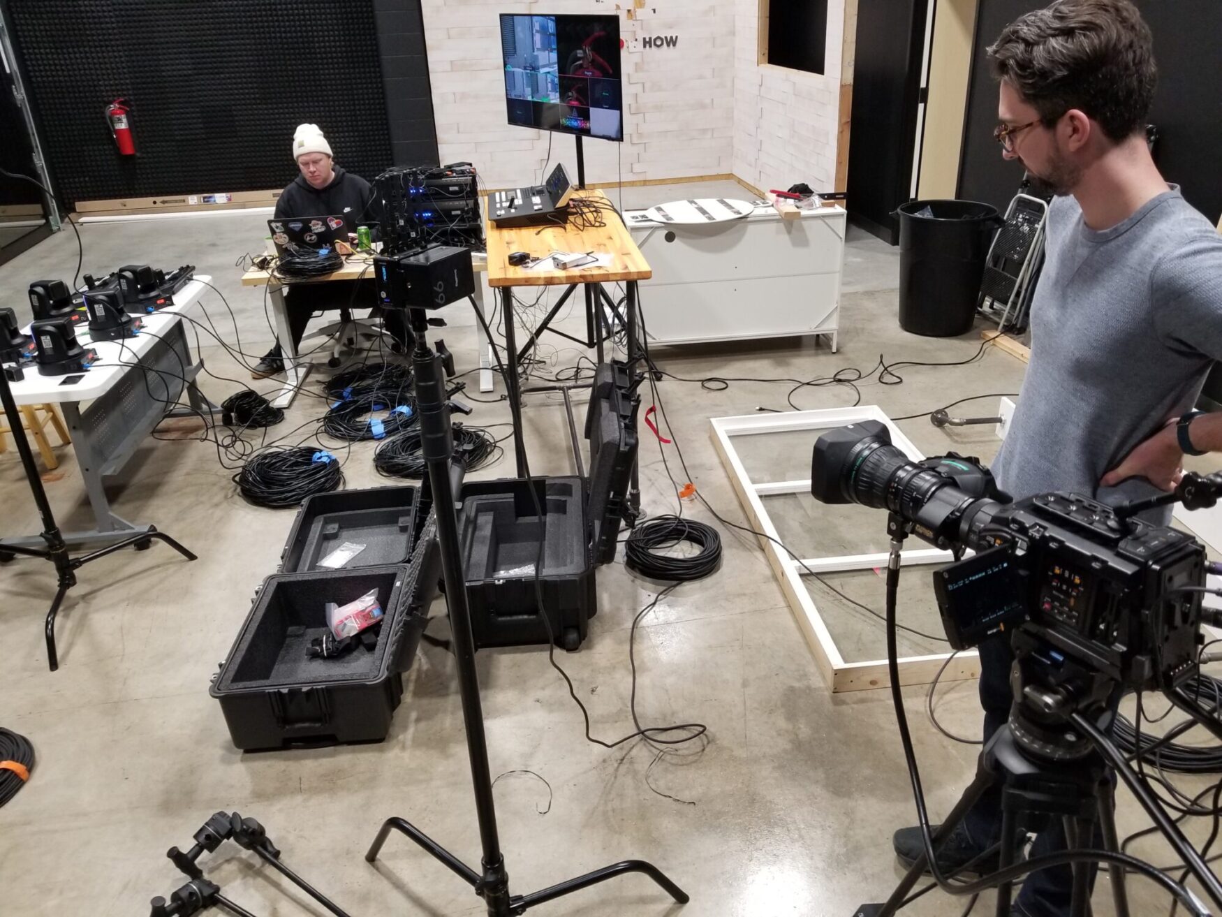 A tech setup scene in a studio with two people. One person sits at a computer with cables and equipment scattered. Another stands near a camera. The vibe is busy and focused.