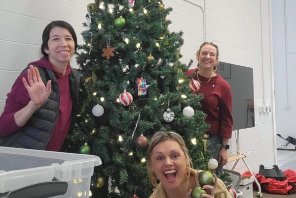 Three people smiling and decorating a Christmas tree with red and white ornaments. One holds baubles, exuding joy and holiday spirit in a festive setting.