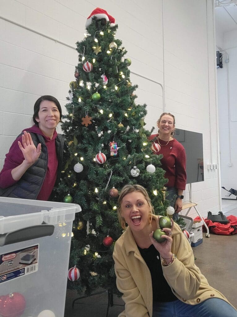Three people smiling and decorating a Christmas tree with red and white ornaments. One holds baubles, exuding joy and holiday spirit in a festive setting.