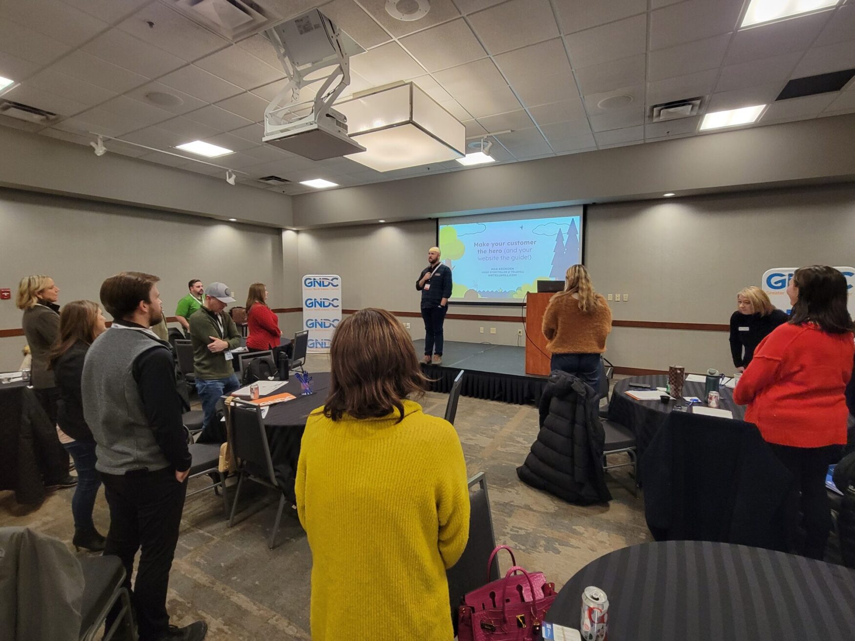 A group of people stands around tables in a conference room, focusing on a speaker on stage. A presentation slide is projected behind him.