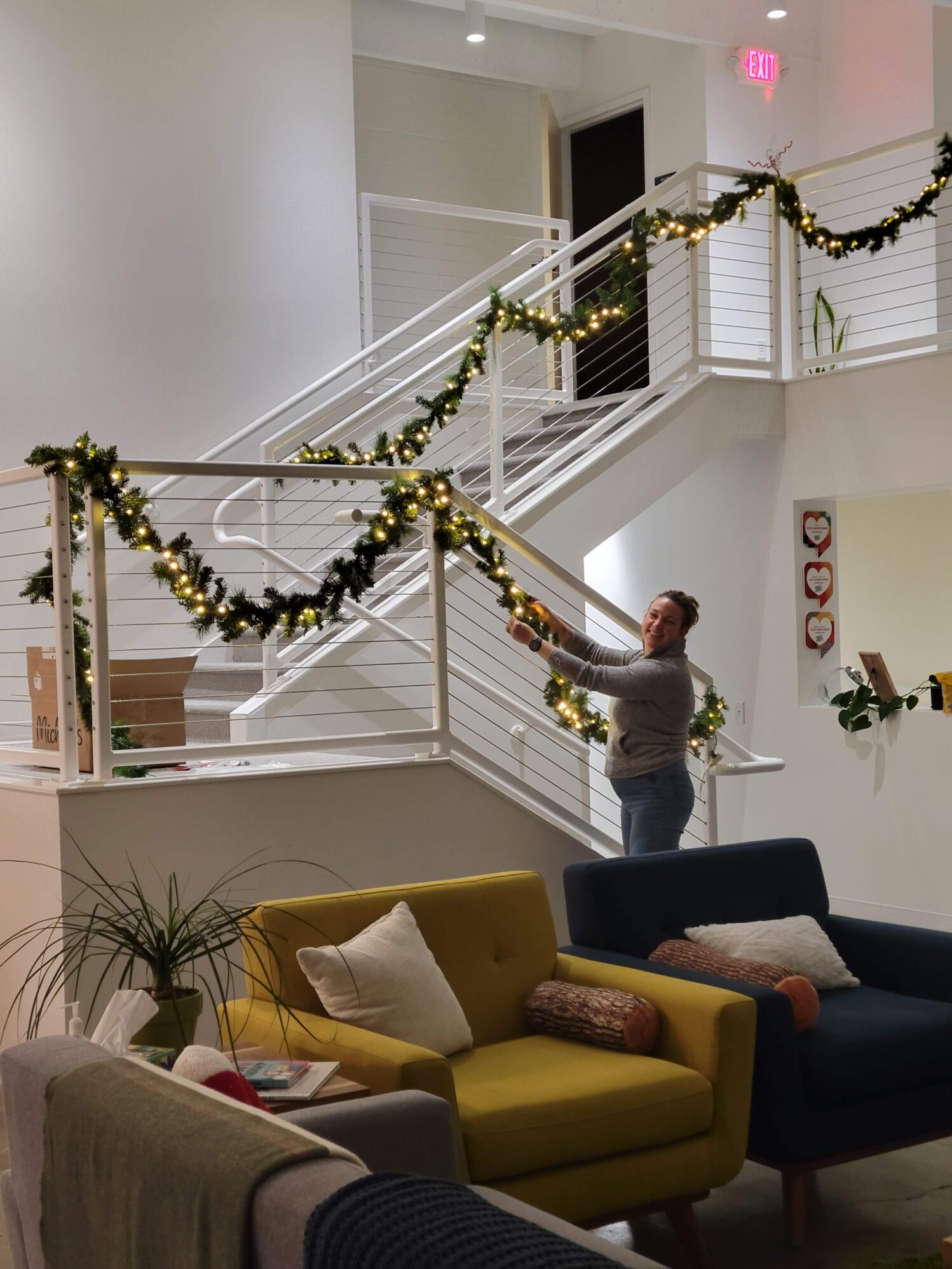 A warmly lit, modern staircase decorated with glowing green garlands. A person joyfully adjusts the decor. Cozy chairs and pillows in the foreground.