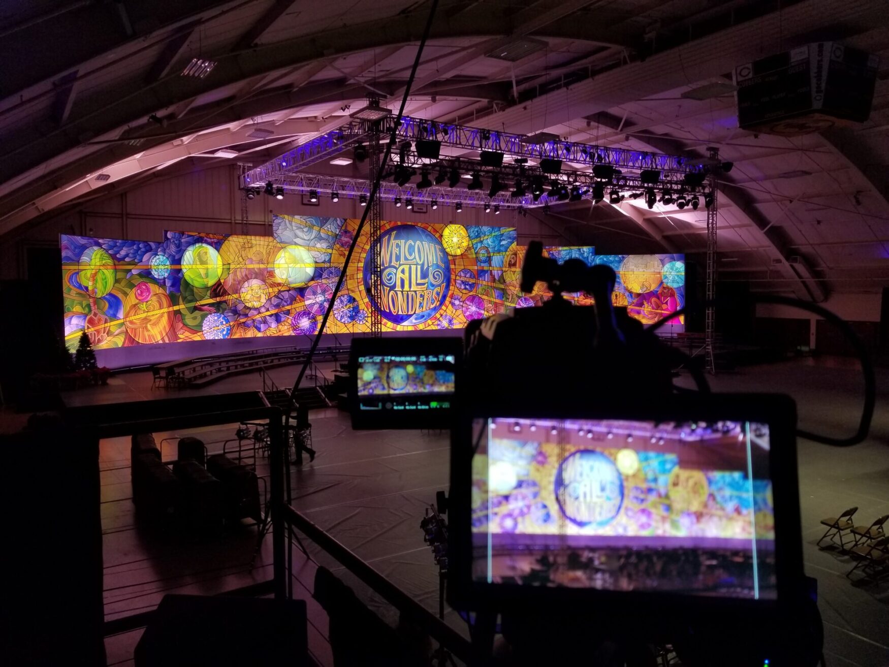 A vibrant stage backdrop displays "Welcome All Wonders" amid colorful abstract designs. Control panels are visible in the foreground. The scene feels festive and welcoming.