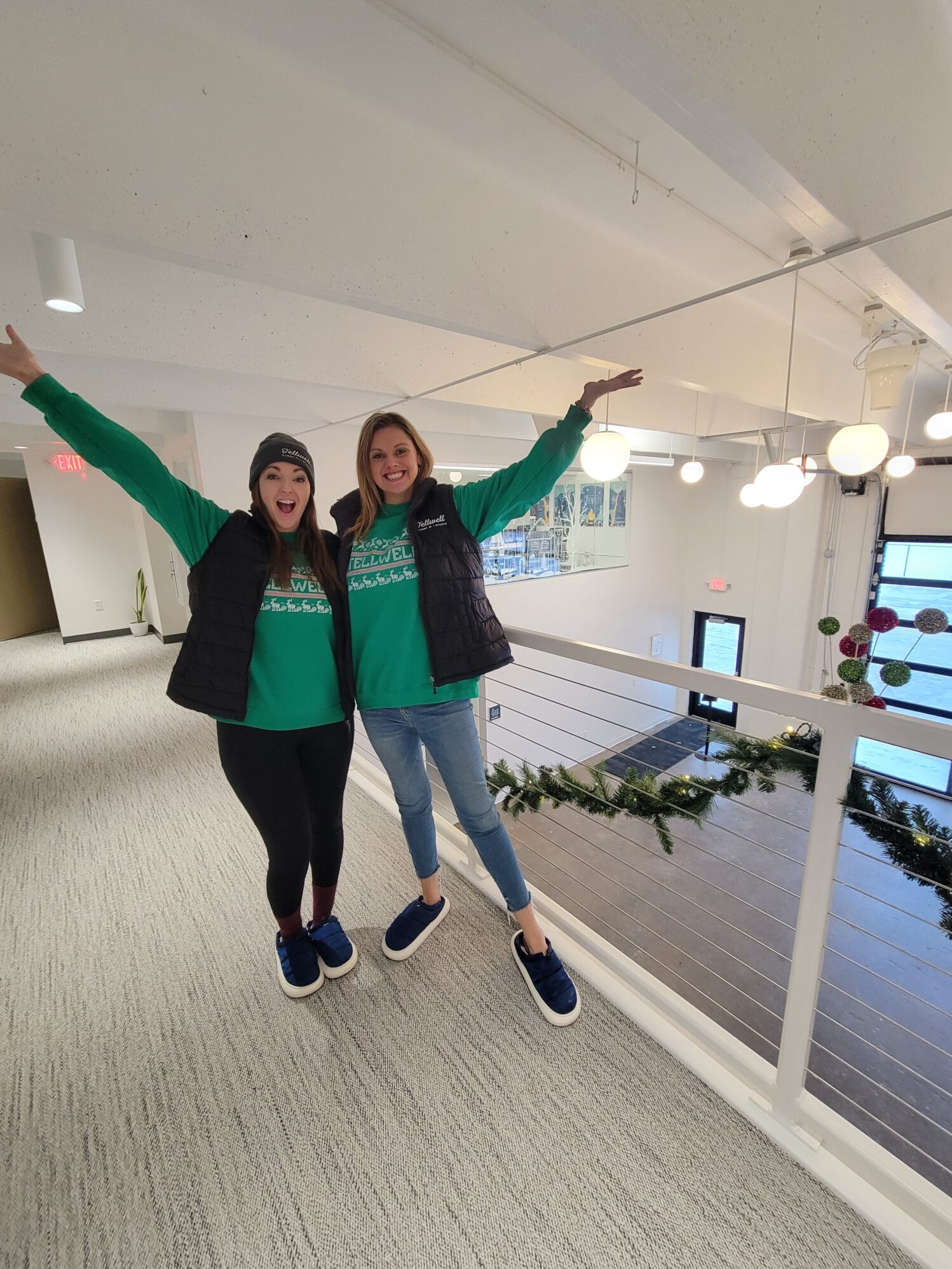 Two women wearing matching green shirts and black vests joyfully pose with arms raised on a decorated indoor balcony. The atmosphere is festive.
