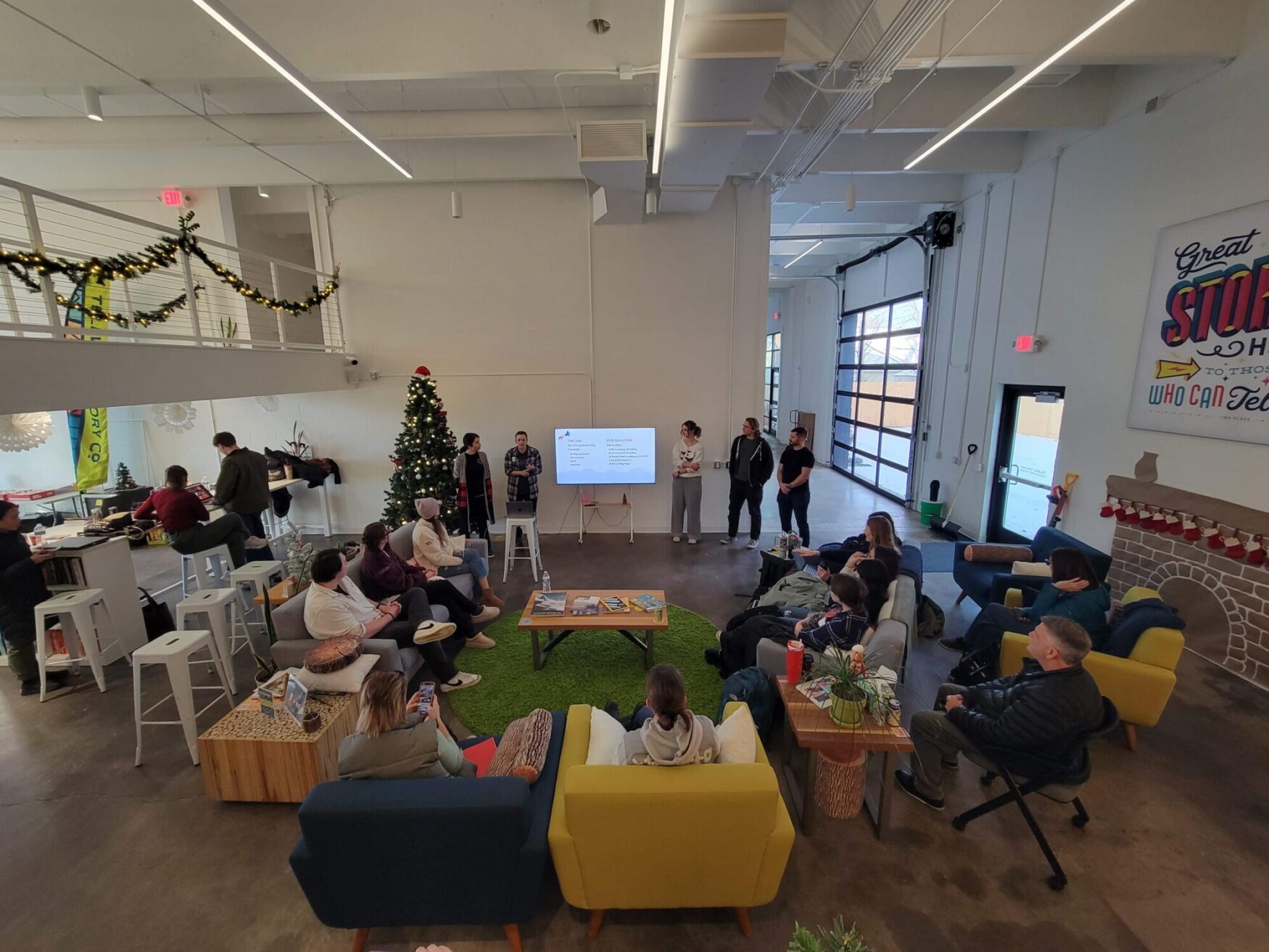 Spacious modern room with people seated on colorful chairs and couches, listening to a presentation. A decorated Christmas tree adds a festive touch.