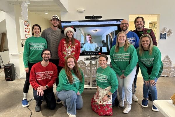 Team members wearing festive Tellwell sweaters pose together in an office during a holiday celebration, smiling in front of a screen showing a remote teammate on a video call.