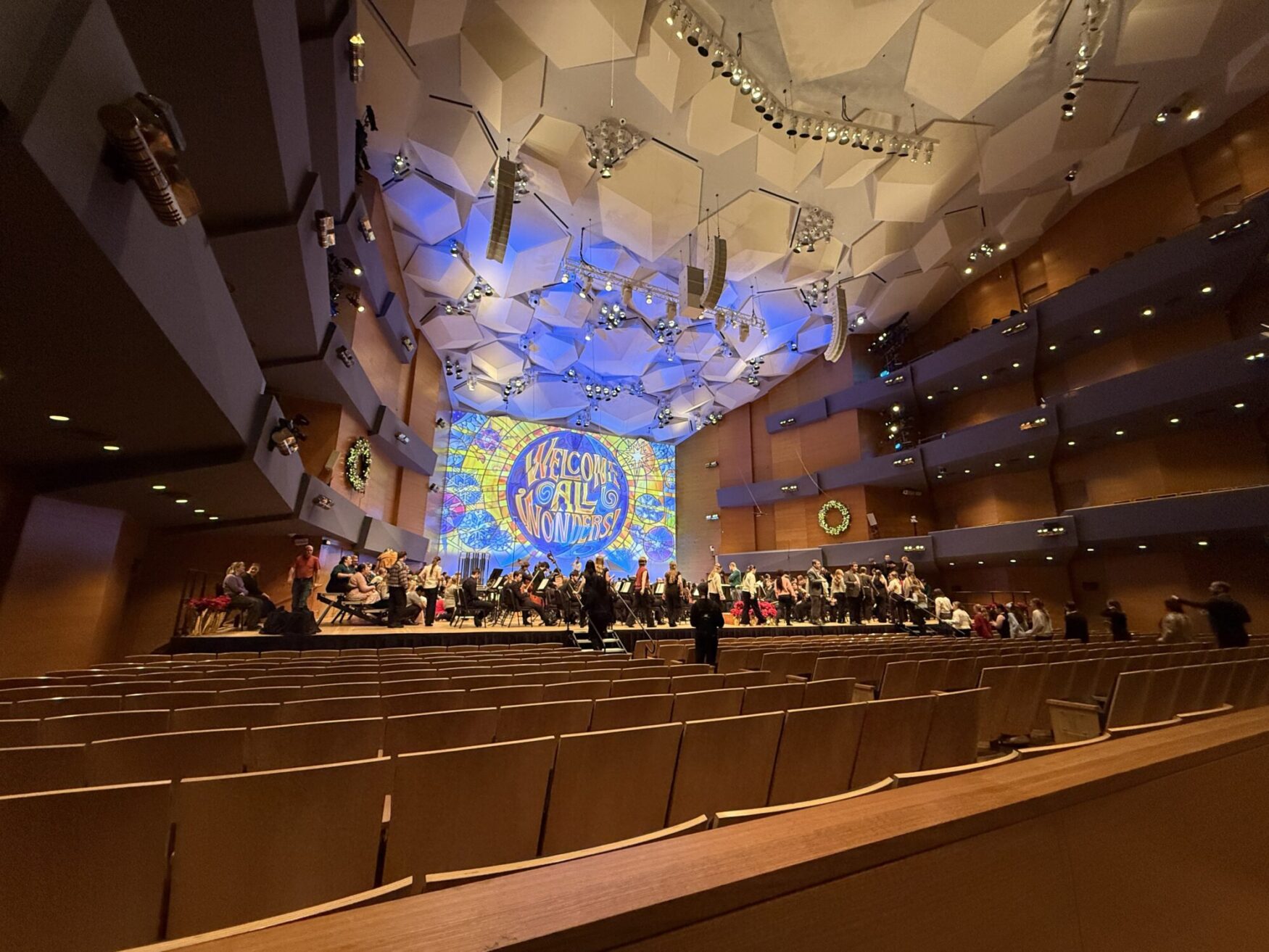 Concert hall with geometric ceiling features a stage with musicians setting up. A bright backdrop reads "Welcome All Wonders," creating a festive atmosphere.
