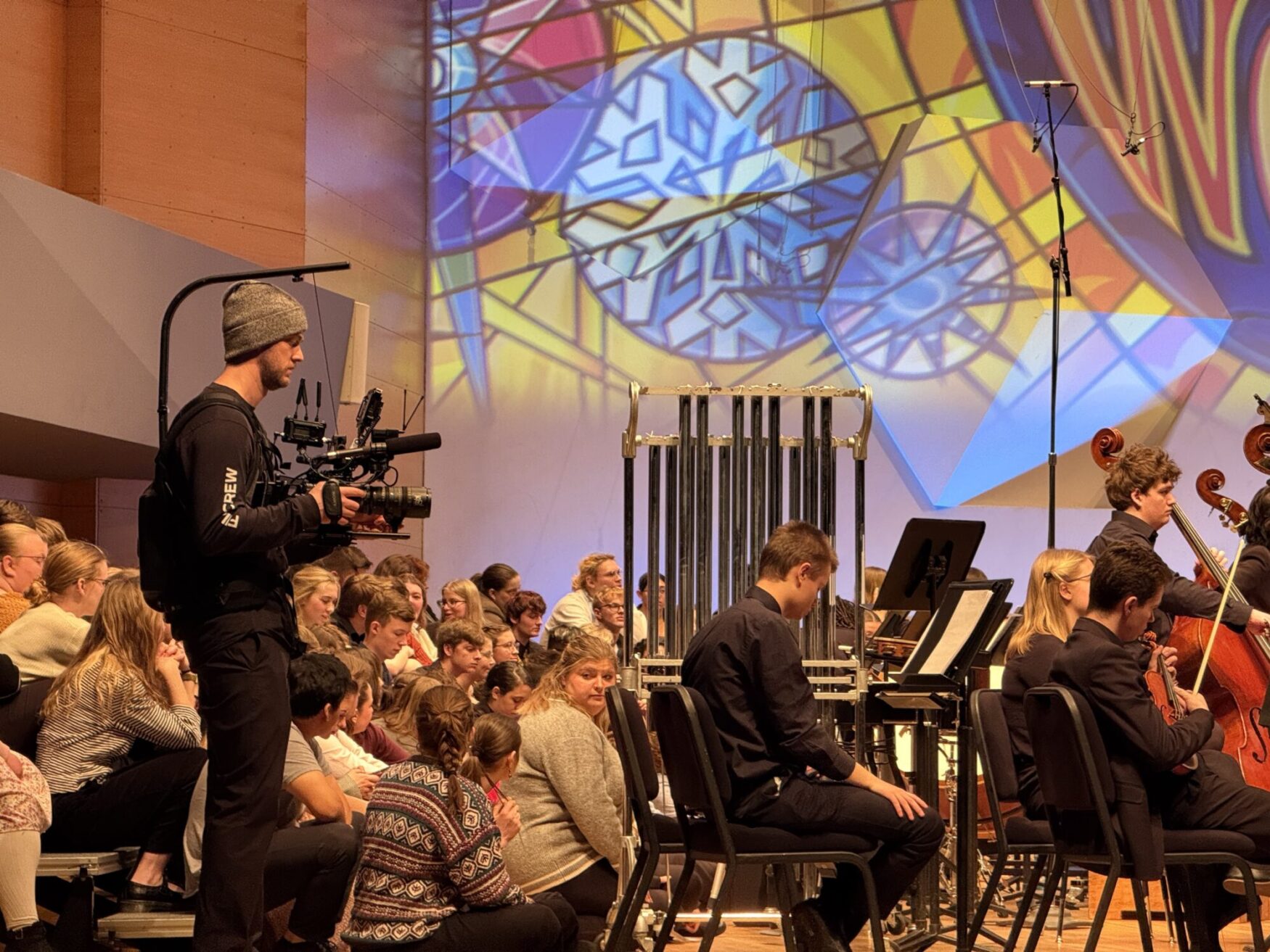 A cameraman in a beanie films an orchestra from the side as colorful stained glass designs are projected on the wall. The audience watches intently.