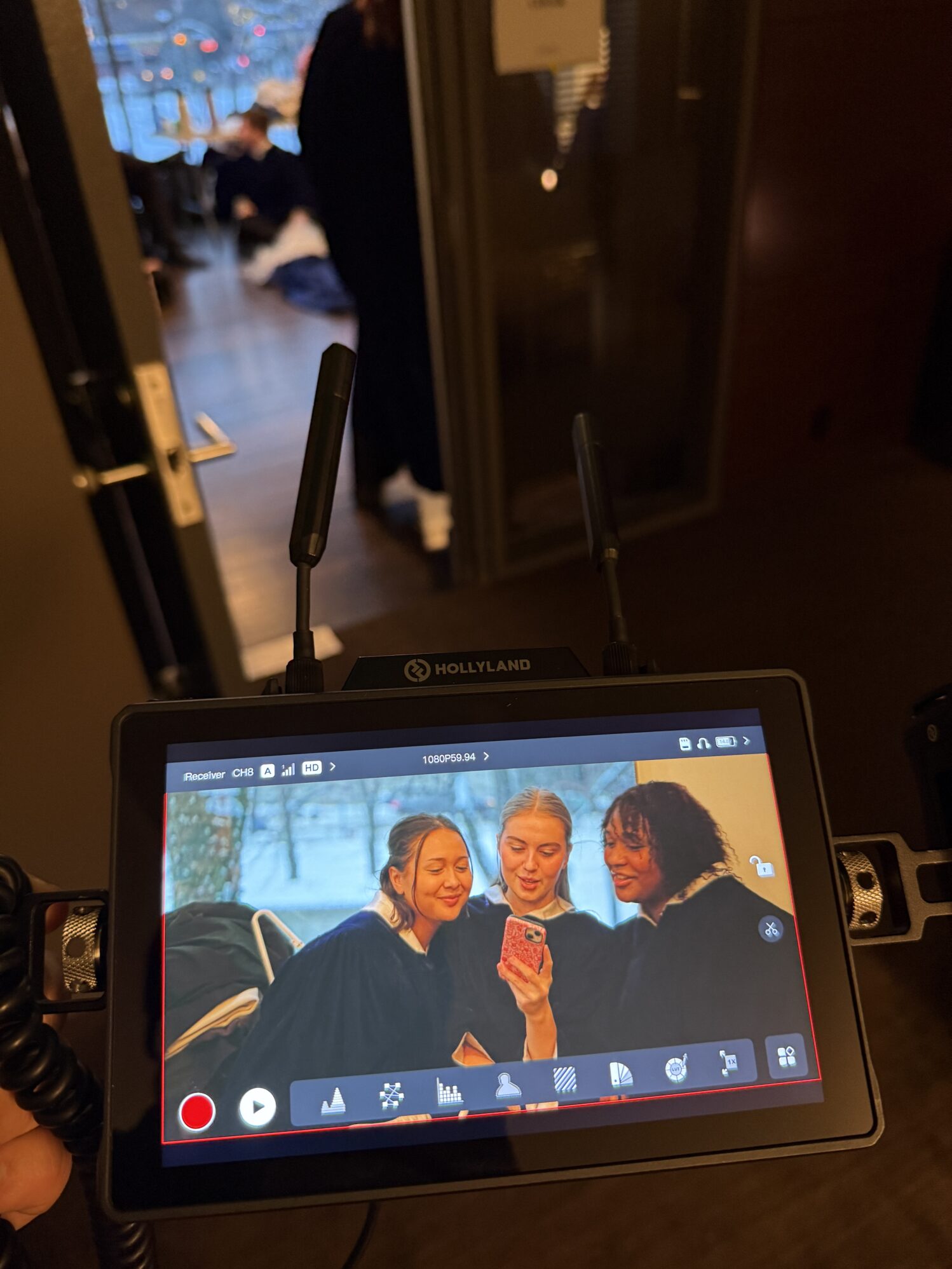 A video monitor shows three people in dark robes joyfully gathered around a phone. In the background, a blurred room with soft lighting and people.