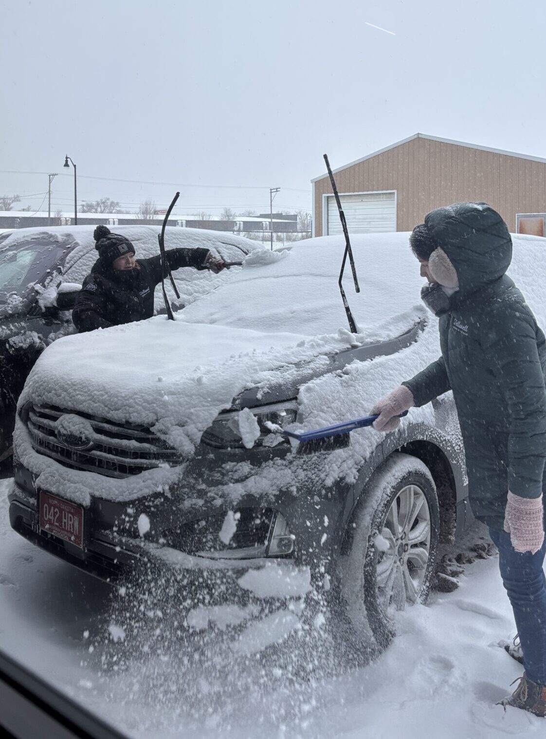 Two people, bundled in winter clothing, clear heavy snow off parked cars. The scene is overcast, and snow covers the ground, creating a chilly atmosphere.