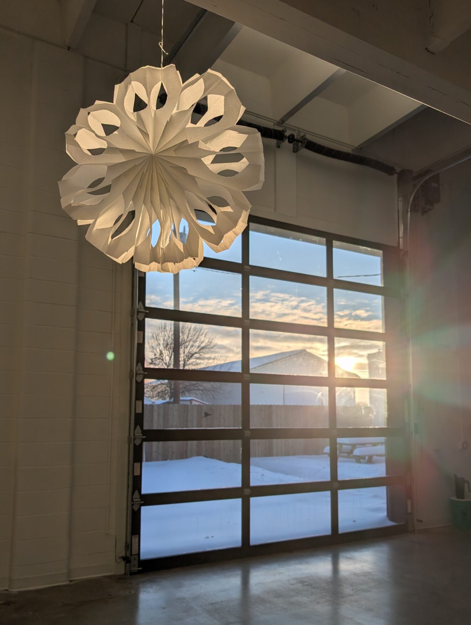 A white paper snowflake decoration hangs in front of a large garage door. Sunlight shines through, casting a warm glow over a snowy outdoor scene.