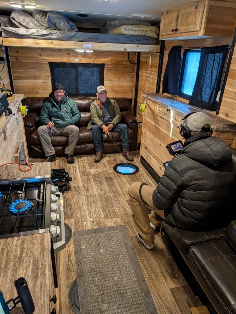 Inside a wood-paneled ice fishing house, a videographer in a black puffer jacket films two men sitting on a leather sofa. The men are smiling and holding fishing rods over a glowing blue hole in the floor, while a gas stove burner glows blue in the foreground.