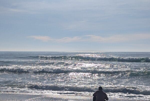 A person in a light-colored shirt crouches on a sandy beach, looking out at the shimmering ocean waves. The sun reflects brightly off the water's surface under a clear, pale blue sky.