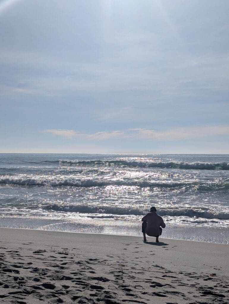 A person in a light-colored shirt crouches on a sandy beach, looking out at the shimmering ocean waves. The sun reflects brightly off the water's surface under a clear, pale blue sky.