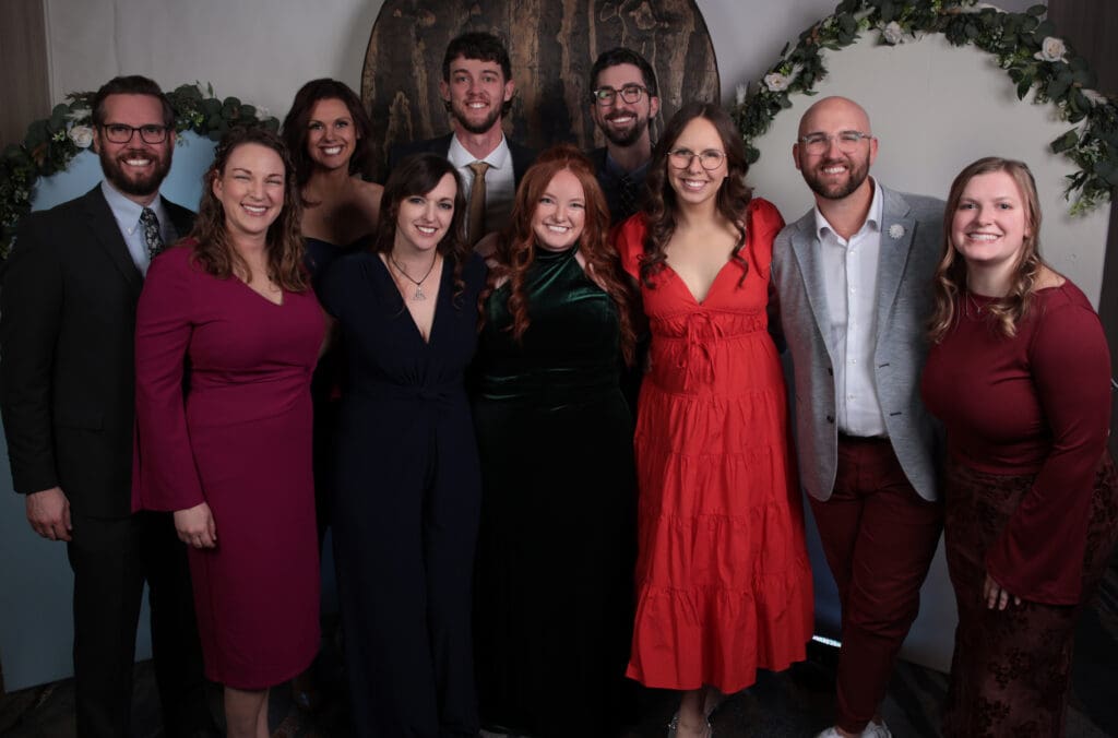 A formal group portrait of ten people dressed in evening wear, posing in front of a photo booth backdrop decorated with a greenery arch. The group is smiling and standing close together.