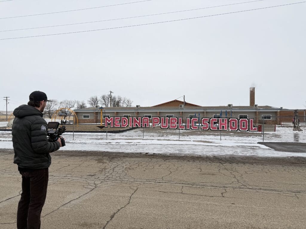A videographer in a puffer jacket films the exterior of Medina Public School. The school's name is displayed in large, red and white block letters on a fence in front of the low-profile brick building on a gray, overcast day.