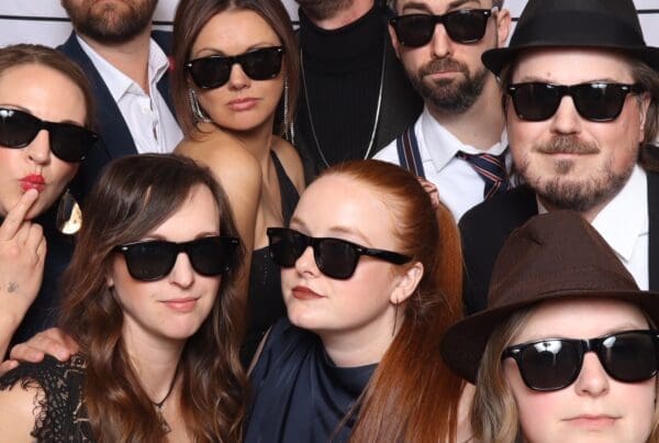 A group photo of nine people posing with serious expressions and wearing matching black sunglasses. They are standing in front of a white backdrop with horizontal black lines, mimicking a police lineup.