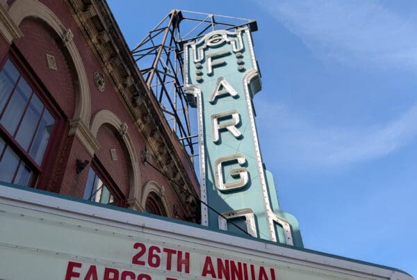 A low-angle shot of the Fargo Theatre’s exterior, showcasing the red brick architecture and the teal-colored vintage neon sign that spells out "FARGO." The marquee below displays "26TH ANNUAL FARGO FILM FESTIVAL" in bold red lettering.