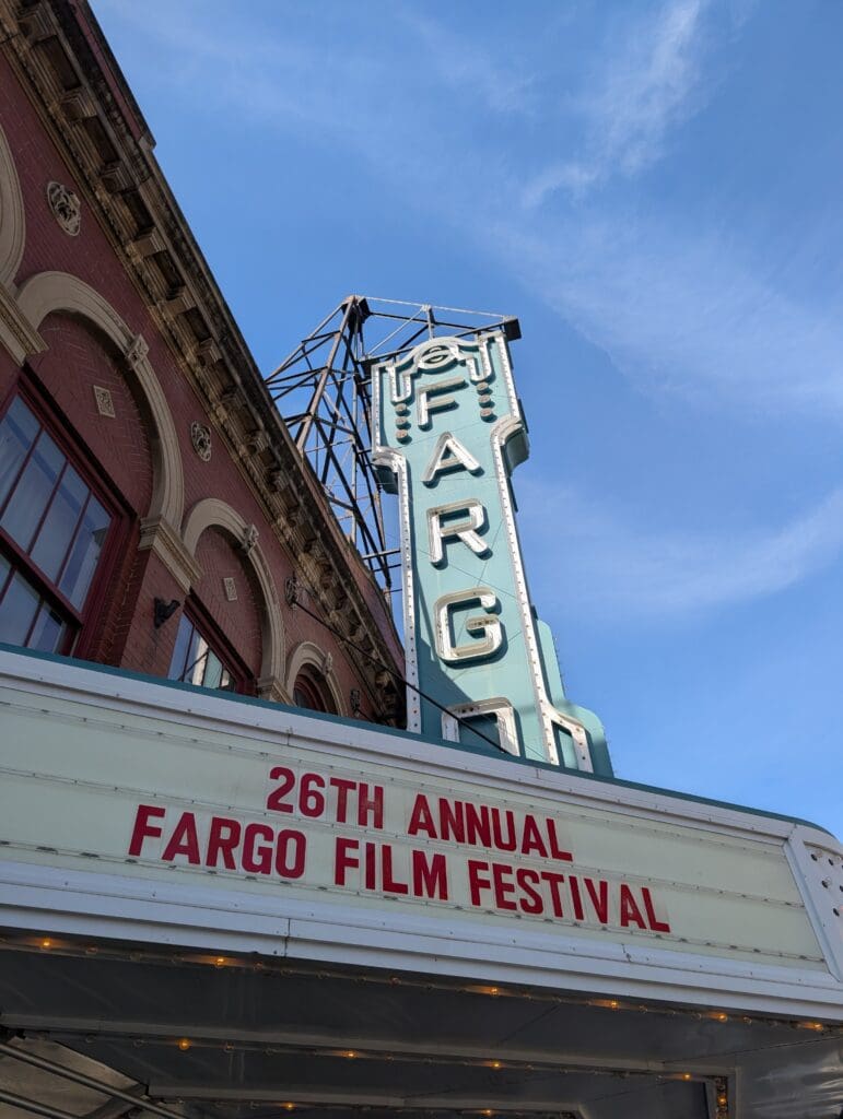 A low-angle shot of the Fargo Theatre’s exterior, showcasing the red brick architecture and the teal-colored vintage neon sign that spells out "FARGO." The marquee below displays "26TH ANNUAL FARGO FILM FESTIVAL" in bold red lettering.