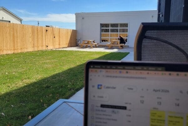 A first-person view from an outdoor table showing a laptop screen with a Google Calendar. In the background, a sunny patio area features a green lawn, a wooden fence, and a person working at a picnic table under a clear blue sky.