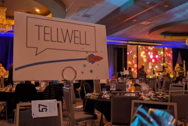A table at a formal event features a place card with the "Tellwell" logo and a rocket ship illustration. In the background, a large ballroom is filled with guests at round tables under colorful stage lighting and a crystal chandelier.