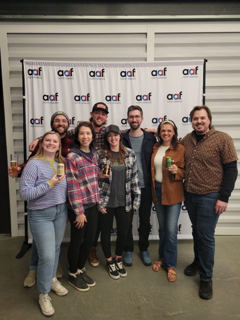 Seven people stand in a group in front of an "AAF North Dakota" branded backdrop. Several team members are smiling and holding up cans of Liquid Death sparkling water and glasses of beer.
