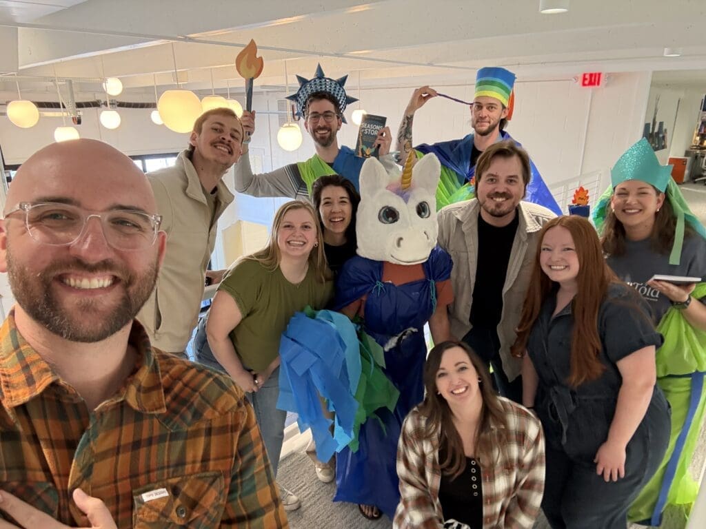 A cheerful group photo of the team in a modern office hallway. Several members are wearing the DIY Statue of Liberty and unicorn costumes made from colorful plastic and tissue paper, while others lean in to join the shot. A man in a plaid shirt takes the selfie in the foreground, capturing everyone’s high energy and smiles.