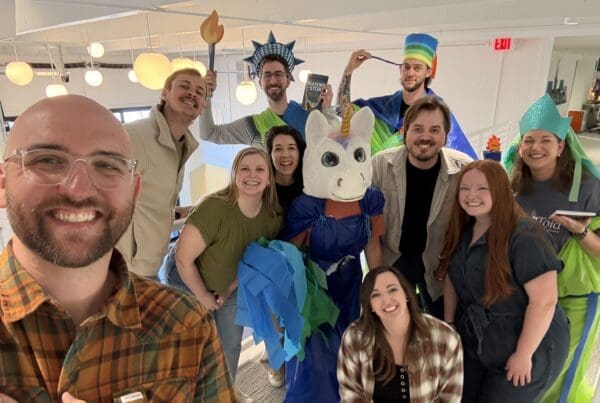 A cheerful group photo of the team in a modern office hallway. Several members are wearing the DIY Statue of Liberty and unicorn costumes made from colorful plastic and tissue paper, while others lean in to join the shot. A man in a plaid shirt takes the selfie in the foreground, capturing everyone’s high energy and smiles.