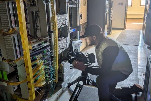 A videographer wearing a baseball cap and safety vest kneels on a walkway, using a tripod-mounted camera to film a complex wall of network server racks. The server room is filled with intricate wiring and glowing indicator lights.