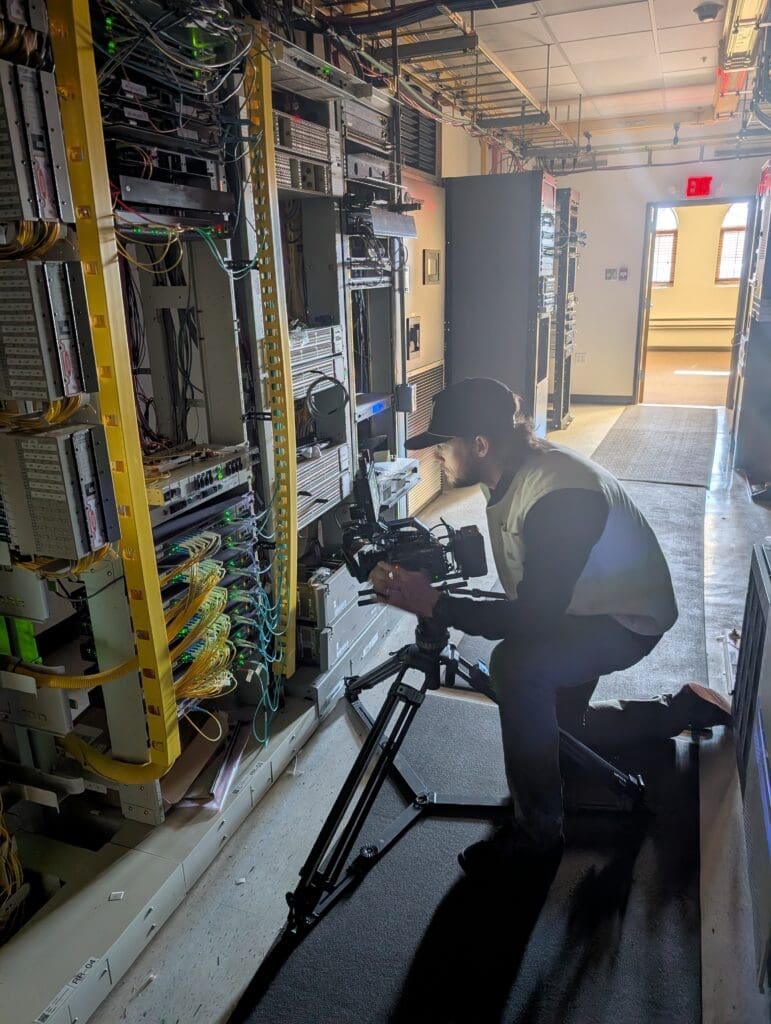A videographer wearing a baseball cap and safety vest kneels on a walkway, using a tripod-mounted camera to film a complex wall of network server racks. The server room is filled with intricate wiring and glowing indicator lights.
