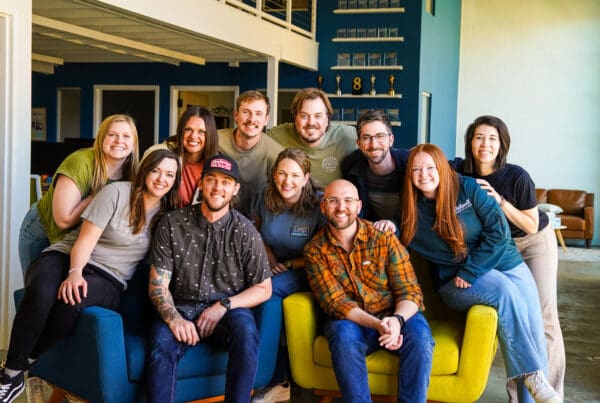 A large group of eleven coworkers are gathered closely together, smiling for a team photo in a modern office space. Two men sit in blue and bright yellow armchairs in the foreground, while the rest of the team leans in behind and around them. A deep blue accent wall with shelves displaying awards is visible in the background.